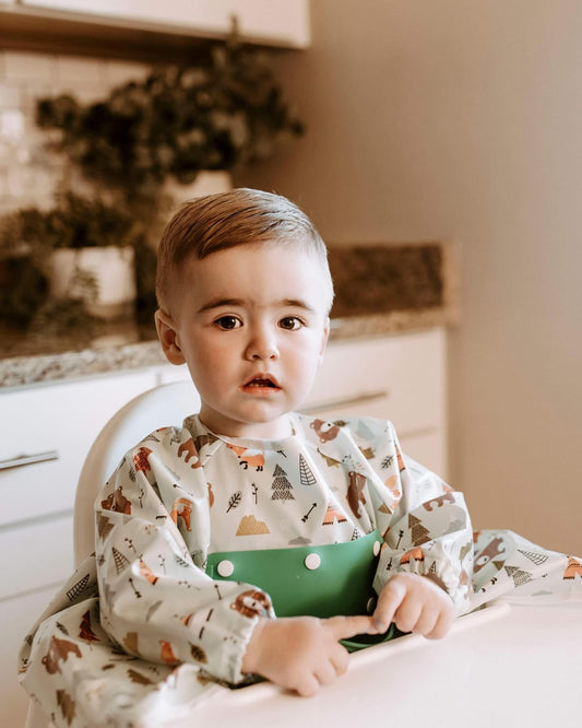 Serene toddler at the table wearing a Bibvy bib with a 'Wilderness Wonder' print, ready for mealtime.