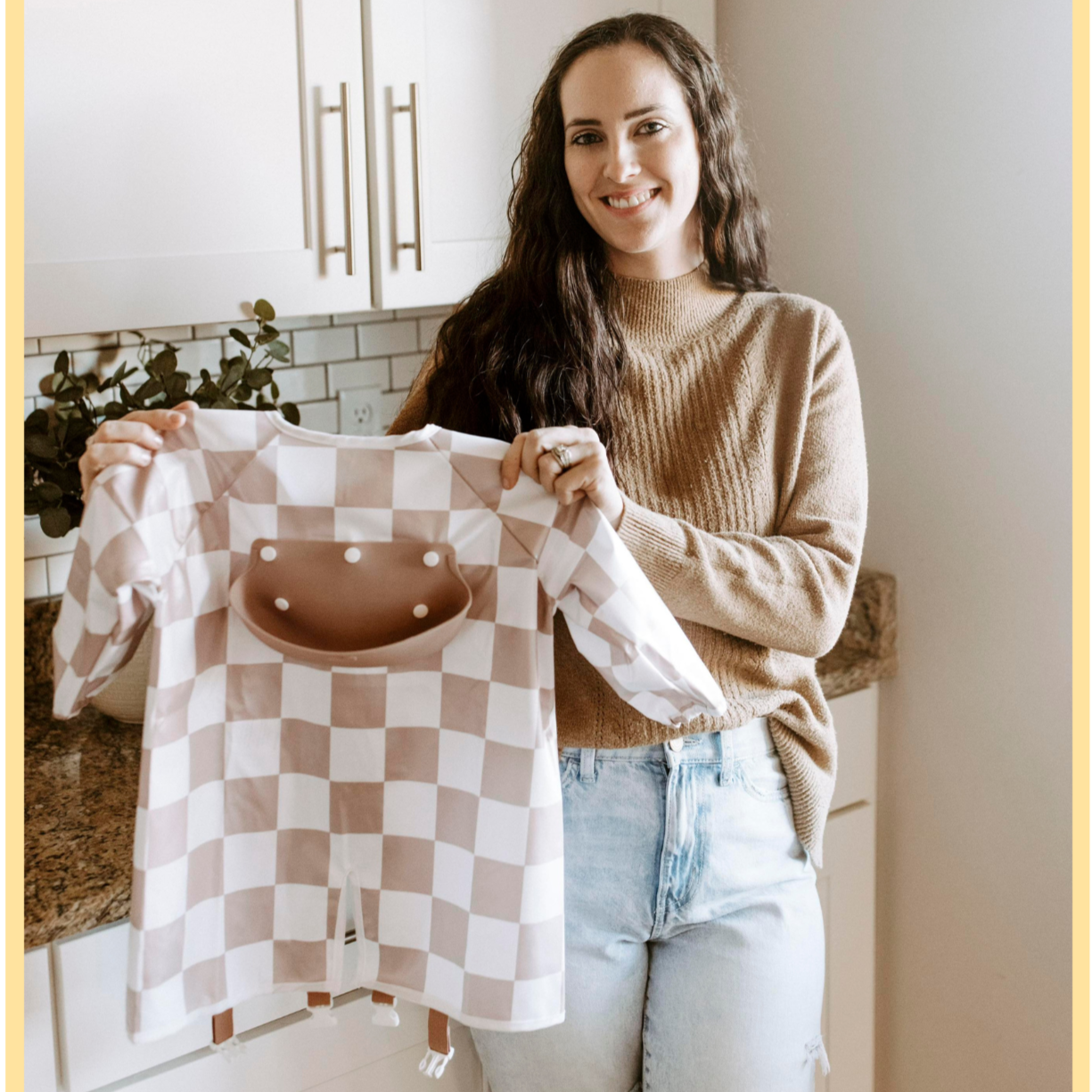 Danielle, the founder of Bibvy, smiling and holding an mauve pink and white checkered bib in a modern kitchen setting.