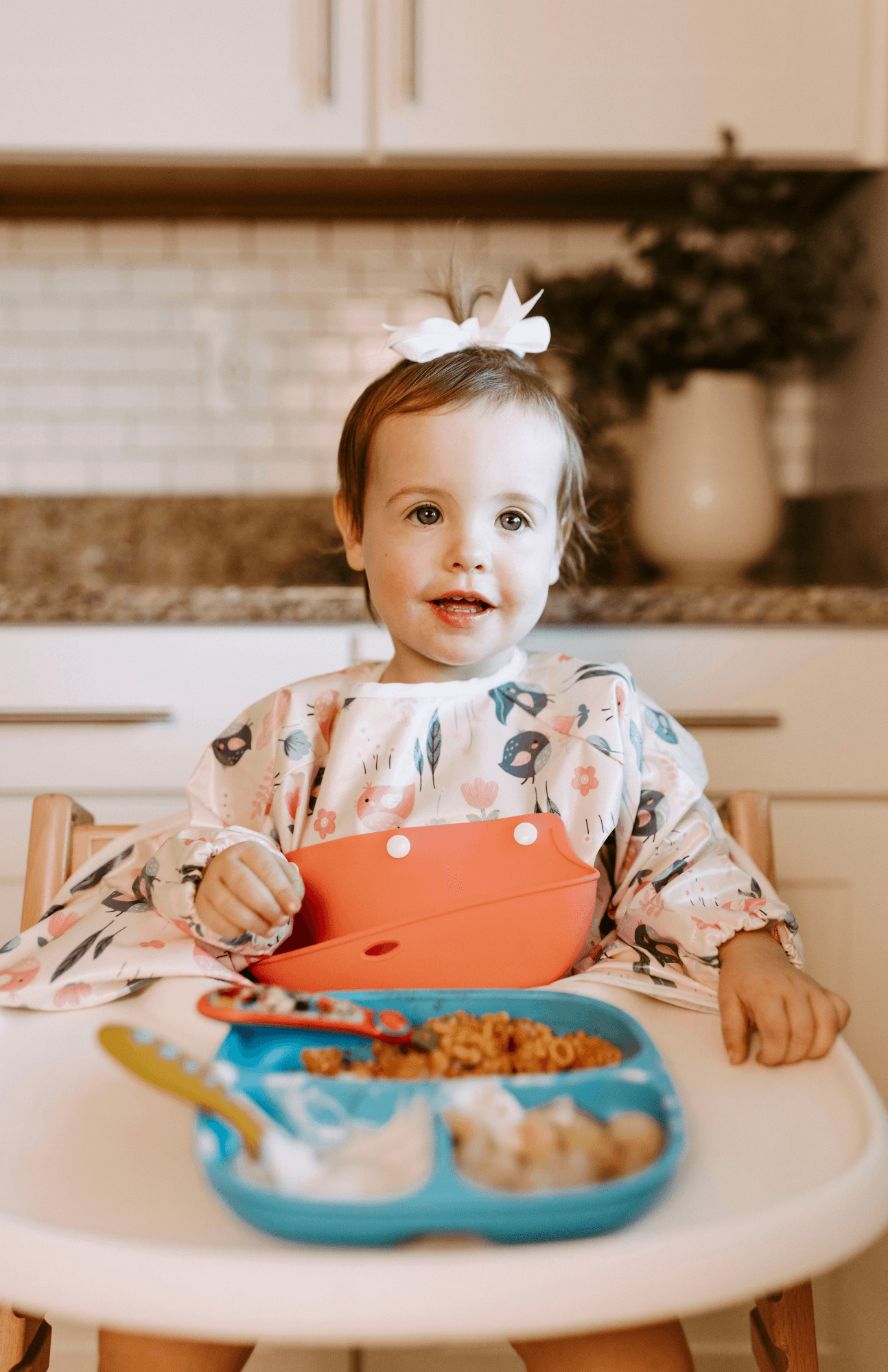 Cheerful baby with a white bow, wearing a Bibvy 'Little Birdie' print bib with a peach silicone food catcher, enjoying a meal in a high chair