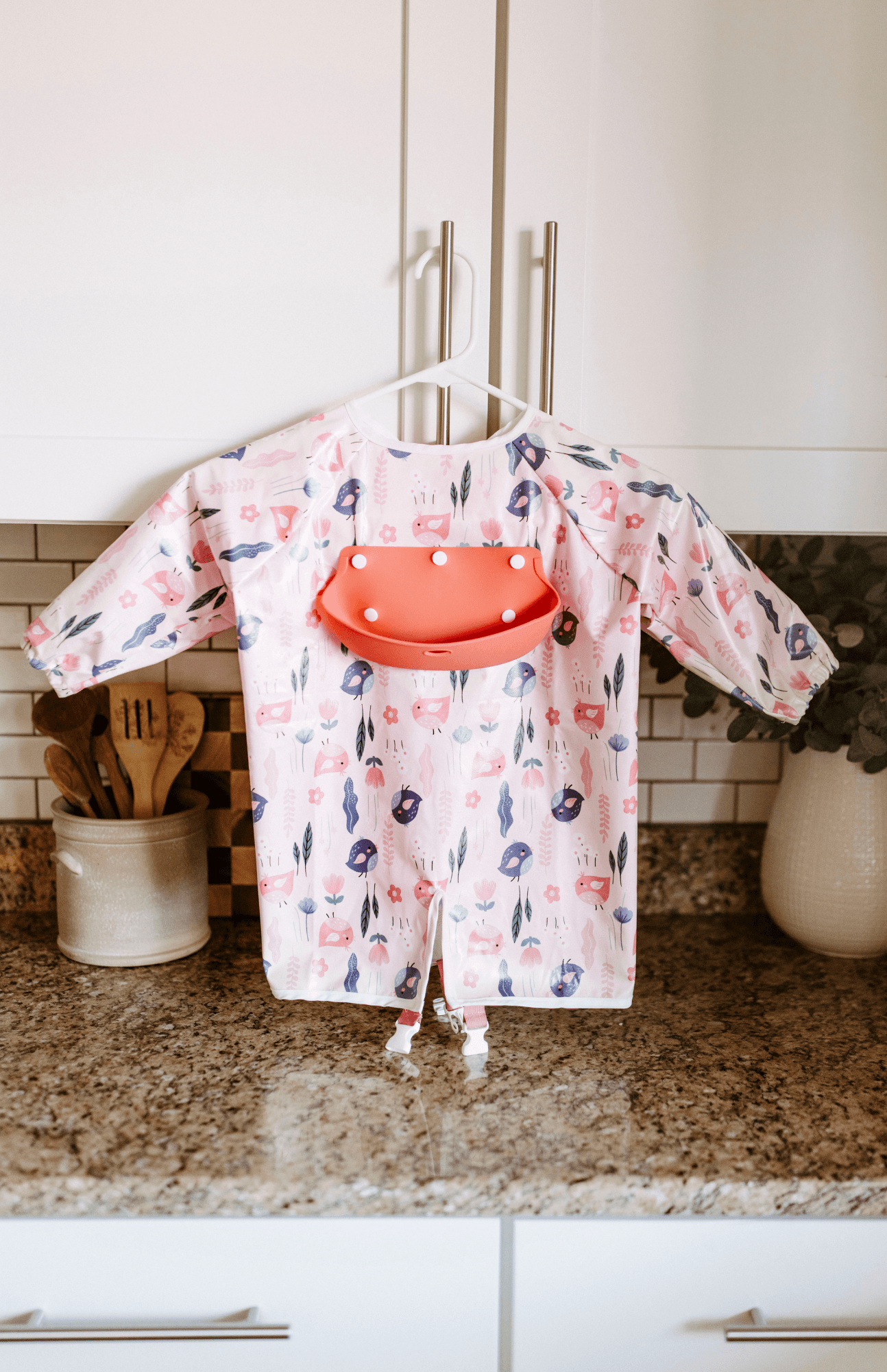 Front view of the Bibvy long sleeve bib in 'Little Birdie' print with a peach silicone food catcher, hanging in a stylish kitchen.