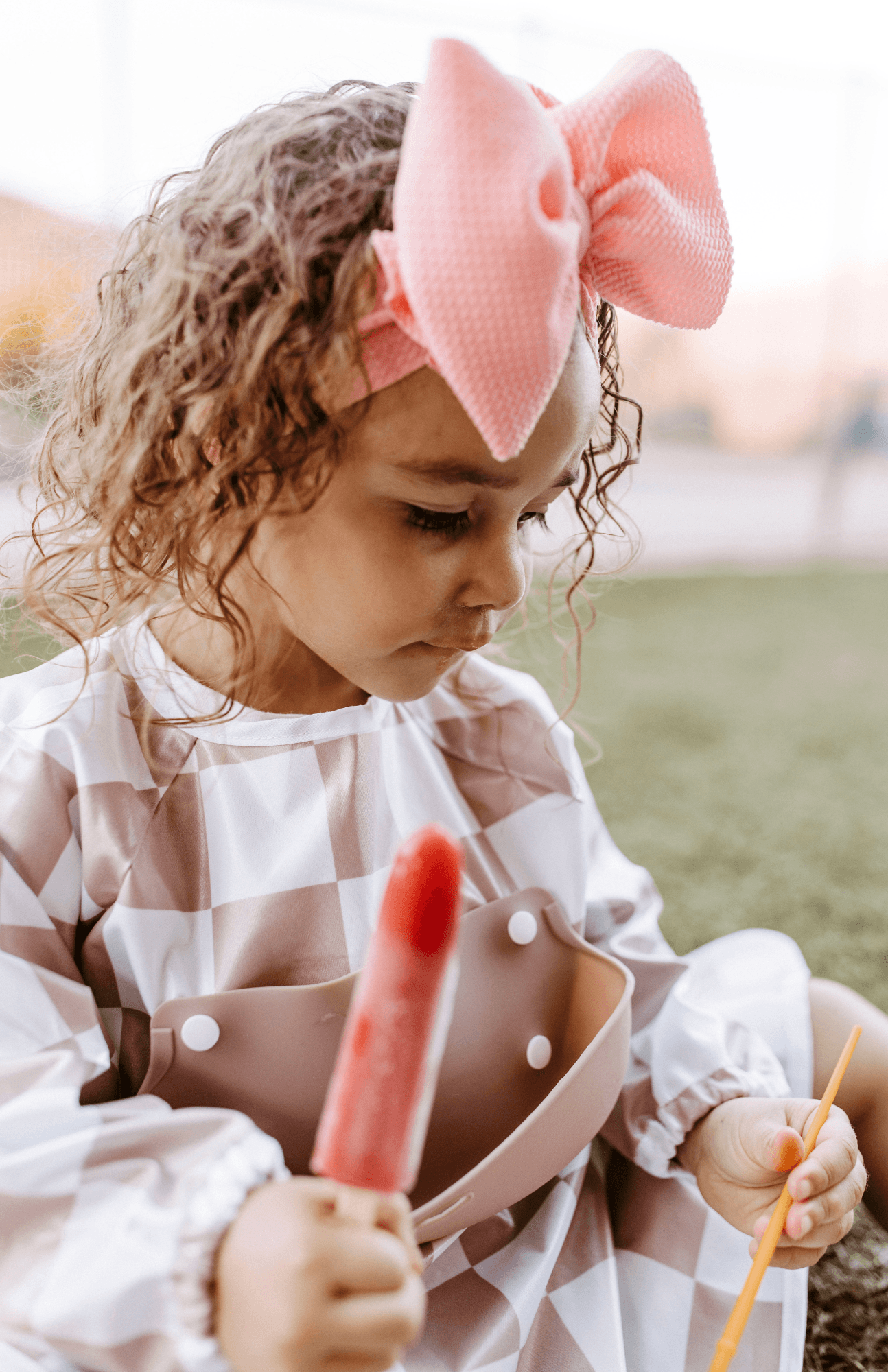 Curly-haired toddler with a pink bow, wearing a Bibvy mauve pink and white checkered smock bib with silicone food catcher, holding a popsicle.