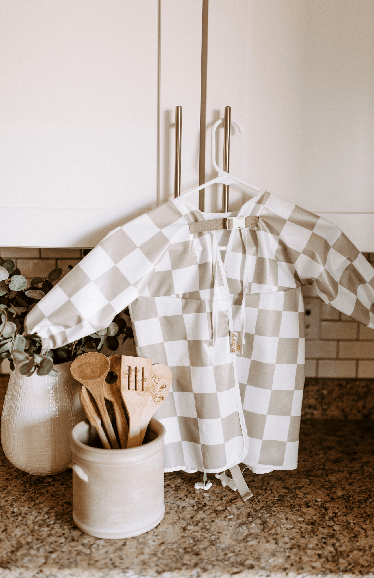 Back view of a olive green and white checkered Bibvy bib hanging in a kitchen, highlighting the tie-up detail for adjustable fit