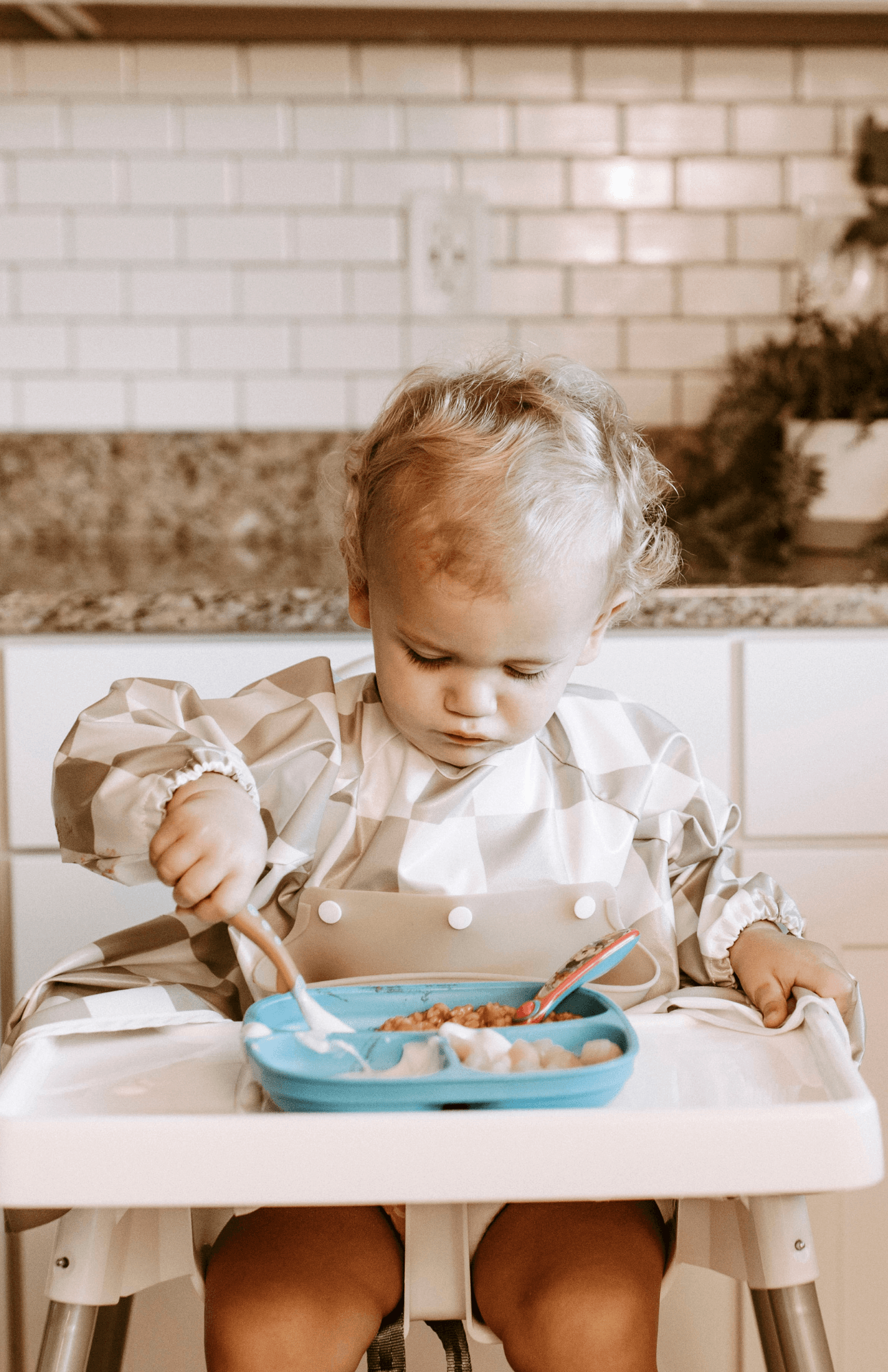 Focused toddler eating from a blue plate while wearing a Bibvy waterproof bib in olive green and white checkered print, seated in a high chair.
